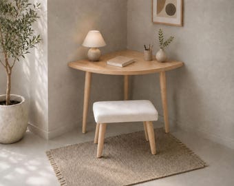 May include: A light-colored wooden corner desk with a curved front edge and three tapered legs. A small white lamp, a closed book, a pencil holder, and a small vase with greenery sit on top. A matching stool with a white upholstered seat is included.