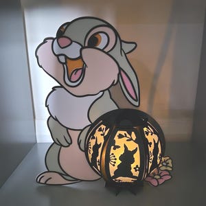 May include: A Bambi-themed decorative set. It features a large, pastel-colored cut-out of the character, alongside a black, spherical lantern with Bambi silhouettes and floral cutouts. The lantern emits a warm glow.