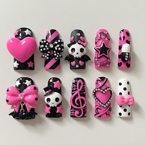 May include: A collection of press-on nails in various designs. The nails feature a black and pink color scheme with embellishments like hearts, bows, skulls, stars, and musical notes. Some nails have a glossy finish, while others have a matte appearance.
