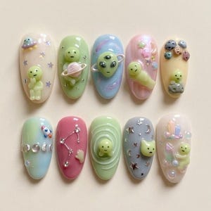 May include: A set of ten artificial nails, each painted in pastel colors and adorned with 3D alien and space-themed designs. The nails feature various embellishments, including stars, planets, and constellations, creating a whimsical and unique aesthetic.