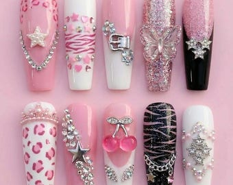 May include: A collection of ten artificial nails in various designs. The nails feature pink, white, and black bases with embellishments like rhinestones, glitter, and charms. Designs include animal prints, stars, hearts, and a butterfly.