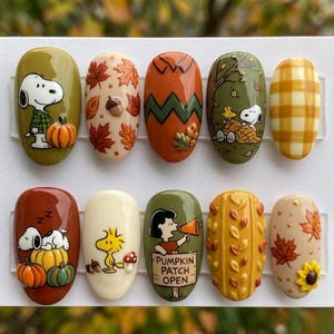 Op de afbeelding: Een set van tien kunstnagels met Peanuts-figuren en herfstthema's. De ontwerpen bevatten Snoopy met een pompoen, esdoornbladeren, een zigzagtruipatroon, Woodstock, ruitjes, een slapende Snoopy en Lucy met een Pumpkin Patch Open-bordje.