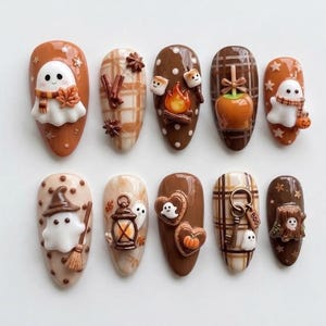 May include: A set of ten press-on nails featuring 3D autumn and Halloween designs. Decorations include ghosts with scarves, a campfire with marshmallows, a caramel apple, a witch hat, a lantern, heart cookies, a key with a tag reading BOO, and a tree stump.