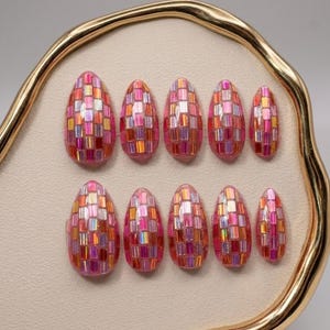 May include: A set of ten almond-shaped press-on nails featuring a mosaic pattern of iridescent pink, orange, and silver rectangular tiles. The nails have a glossy finish and are arranged in two rows on a cream-coloured display tray with a gold rim.