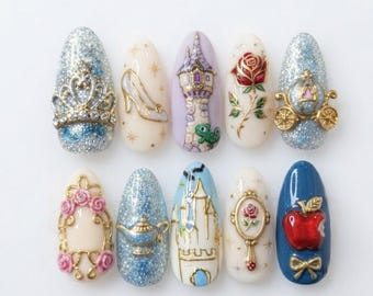 Peut inclure: Un ensemble de dix faux ongles en forme d'amande avec des motifs 3D complexes, notamment une couronne dorée, un soulier de verre, une tour, une rose rouge, un carrosse doré, un cadre floral, une lampe magique, un château, un miroir et une pomme.