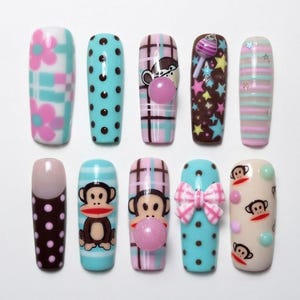 May include: A set of ten press-on nails featuring various designs including pink flowers on a plaid background, black polka dots on light blue, a monkey face with a pink bubble, colourful stars on brown, stripes, a bow, and monkey patterns.