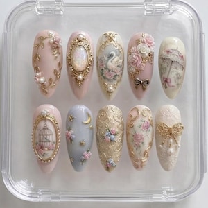 May include: A set of ten almond-shaped press-on nails arranged in a clear plastic case. The designs feature intricate 3D gold filigree, miniature pearls, hand-painted floral patterns, a swan, a birdcage, a parasol, and small metallic bows.