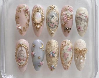 May include: A set of ten almond-shaped press-on nails arranged in a clear plastic case. The designs feature intricate 3D gold filigree, miniature pearls, hand-painted floral patterns, a swan, a birdcage, a parasol, and small metallic bows.