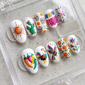 Peut inclure: Un ensemble de dix faux ongles blancs disposés dans un boîtier en plastique transparent. Chaque ongle présente des motifs peints à la main, notamment un soleil, un cactus, des crânes, des fleurs, un coq, un oiseau et des piments aux couleurs vives.