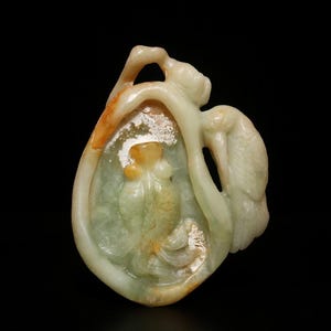 May include: A carved jade pendant in shades of green, cream, and orange. The pendant features a central figure within a teardrop shape, with a bird-like form on the side. The jade is translucent, with intricate details.