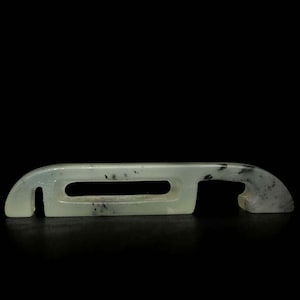 May include: A pale green jade artifact with a rectangular central opening and curved ends. The jade has dark speckling and a smooth, polished surface. The object is set against a black background.