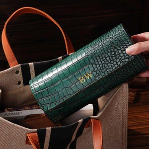 May include: A green, crocodile-textured clutch with gold initials "BW" is held near a canvas tote bag with black stripes and brown leather handles. The bag contains books and a small brown pouch. The background is a dark wood surface.
