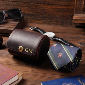 Può includere: Un astuccio per orologio in pelle marrone con le iniziali "GM" in lettere dorate, una cravatta arrotolata, un passaporto e un orologio su una superficie di legno. Sono visibili anche occhiali da sole e libri.