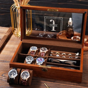 Puede incluir: Una caja de madera para relojes y joyas con tapa de cristal. La caja contiene varios relojes con correas de cuero, anillos y un par de gafas. También se ven una botella pequeña y un jarrón decorativo.