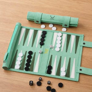 May include: A mint green fabric backgammon set featuring dark olive and white triangular points. The board includes white and black circular checkers, two standard dice, and a doubling cube with the number 64. A matching roll-up case displays the name JAMES.