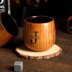 May include: A wooden tumbler with a dark interior and a natural wood grain exterior. The tumbler features the engraved initial "J" with the name "JAMES" below it. It sits on a small wooden coaster. Beside it are two grey square ice cubes.