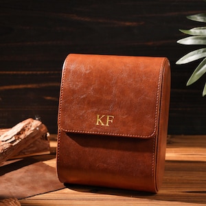 May include: A brown leather watch case with a flap closure. The initials "KF" are embossed in gold on the front. The case is rectangular and sits on a wooden surface.