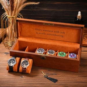 May include: A rectangular wooden watch box with a hinged lid, lined in orange velvet, featuring the text "Every moment with you is worth remembering." The box contains four watches inside and two watches on orange cushions in front, with a small metal key.
