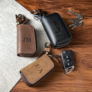 May include: Three leather key cases with metal zips and key rings. One case is smooth brown leather with the initials JM, one is textured black leather with the initial J, and one is light brown leather with the initials JA. A car key fob is included.