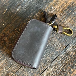 Pode incluir: Um estojo de chaves em couro castanho escuro com acabamento liso e costuras vis&iacute;veis ao longo das bordas. O estojo possui um fecho de correr envolvente e um gancho girat&oacute;rio de metal em tom de lat&atilde;o antigo para prender as chaves.