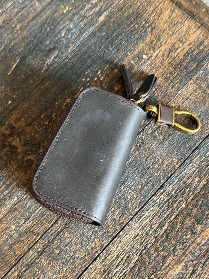 Pode incluir: Um estojo de chaves em couro castanho escuro com acabamento liso e costuras vis&iacute;veis ao longo das bordas. O estojo possui um fecho de correr envolvente e um gancho girat&oacute;rio de metal em tom de lat&atilde;o antigo para prender as chaves.