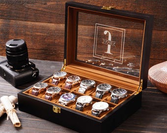 May include: A black wooden watch display case with a clear glass top, holding ten wristwatches with various band and face designs. The interior is lined with a tan fabric. The glass top has the word "JAMES" etched on it.