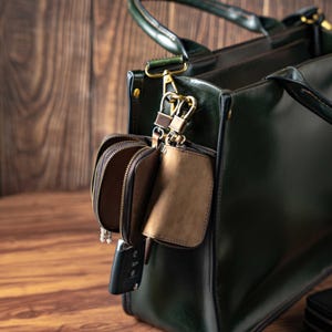 Pode incluir: Uma bolsa tote de couro verde floresta escuro com duas bolsas de couro marrom com z&iacute;per anexadas. As bolsas s&atilde;o presas &agrave; bolsa com fechos girat&oacute;rios de metal cor de lat&atilde;o. Uma chave de carro preta est&aacute; pendurada no z&iacute;per de uma bolsa.