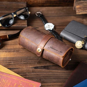 Könnte beinhalten: Eine braune Leder-Uhrenrolle mit Riemenverschluss und dem eingravierten Namen "James Su". Die Rolle ist umgeben von einer schwarzen Uhr, Sonnenbrille, Pfeife und einem kleinen Portemonnaie auf einer Holzoberfläche.