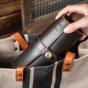 Könnte beinhalten: Eine dunkelbraune Leder-Rolltasche mit den Initialen "JIA" auf der Vorderseite. Die Tasche wird mit einem passenden Lederriemen und einem messingfarbenen Druckknopfverschluss gesichert. Die Tasche befindet sich in einer gestreiften Tragetasche mit braunen Ledergriffen.