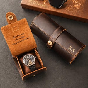 Könnte beinhalten: Eine dunkelbraune Uhrenrolle aus Leder mit den eingeprägten Initialen KA auf der Vorderseite und einem Druckknopfverschluss. Ein offenes Uhrenetui zeigt eine Armbanduhr, mit dem eingravierten Text For the man who makes every moment count.