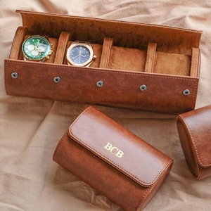 Puede incluir: Un estuche de reloj de cuero marrón con dos relojes en el interior. Los relojes tienen esferas verdes y azules. También se ve un estuche de reloj de cuero marrón más pequeño con las iniciales "BCB".