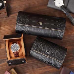 May include: Black leather watch cases with a crocodile texture, featuring gold initials. One case is open, displaying a watch with a brown leather band. A second watch with a black band is also visible. The cases are on a wooden surface.