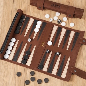 Può includere: Un set da backgammon marrone con pedine bianche e nere, dadi e una custodia. Il tabellone del backgammon è aperto, mostrando la superficie di gioco con triangoli neri, beige e bianchi. La custodia è marrone e ha il nome "JAMES" stampato sopra.