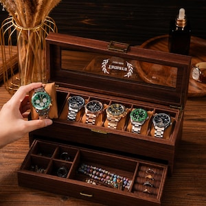 May include: A dark wood watch box with a glass lid featuring a white laurel wreath logo and the text "ETIMET". The box includes a top compartment with six watch pillows and a pull-out drawer with multiple storage sections for jewelry.