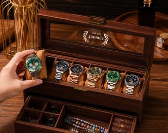 May include: A dark wood watch box with a glass lid featuring a white laurel wreath logo and the text "ETIMET". The box includes a top compartment with six watch pillows and a pull-out drawer with multiple storage sections for jewelry.