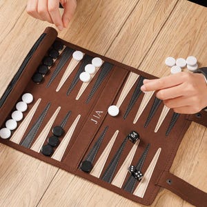 May include: A brown roll-up backgammon board featuring alternating black and beige triangular points. The board includes white and black circular checkers, two black dice, and one doubling cube marked with the number 64. The text JIA is printed in white on the center of the board.