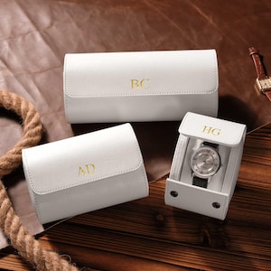 Puede incluir: Tres estuches de reloj de cuero blanco con iniciales doradas. Un estuche está abierto, mostrando un reloj plateado con una correa negra. Las iniciales "BC", "AD" y "HG" están grabadas. Los estuches están sobre una superficie de madera.