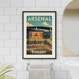 May include: A framed art print featuring Arsenal's Emirates Stadium. The artwork has a vintage travel poster style, with the word "ARSENAL" at the top. The stadium is depicted in detail, with a crowd of people and the Emirates logo.