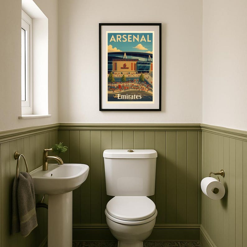 May include: A framed poster with the text "ARSENAL Emirates" hangs above a white toilet in a bathroom. The poster features a stadium illustration with a blue sky background. The bathroom has a white sink, gold fixtures, and olive green wainscoting.