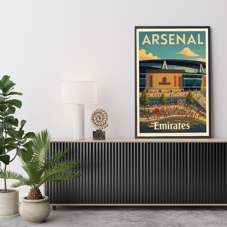 May include: A framed poster featuring the Emirates Stadium, home of Arsenal Football Club. The artwork has a vintage travel poster style, with the word "ARSENAL" at the top and "Emirates" at the bottom. The stadium is depicted with a red and beige color scheme.