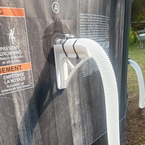 Könnte beinhalten: Nahaufnahme eines Poolabschnitts mit Holzoptik. Eine weiße Kunststoffhalterung stützt einen weißen, geriffelten Schlauch. Das Warnschild lautet: "PREVENT DROWNING Watch children at all times."
