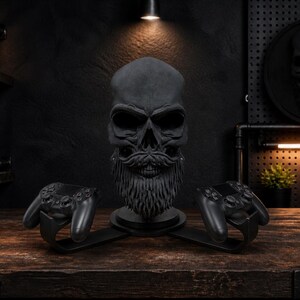 Könnte beinhalten: Eine mattschwarze Totenkopfskulptur mit detailliertem Schnurrbart und Bart, die als zentraler Ständer dient. Zwei schwarze Gaming-Controller sind auf geschwungenen Armen positioniert, die von der Basis des Schädels ausgehen und eine Einheit bilden.