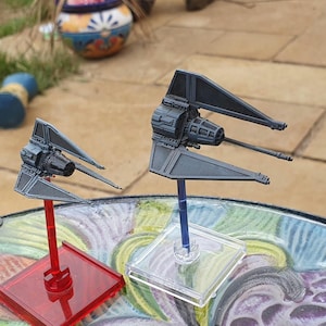 May include: Two gray model starfighters with angled solar panels and detailed cockpits are displayed on clear and colored stands. One is on a red stand, the other on a clear stand. The models are set against a blurred outdoor background.
