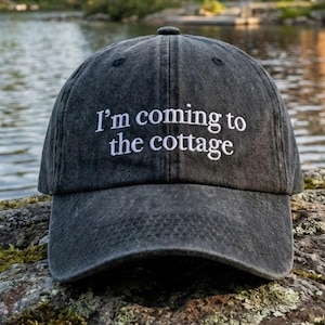Pode incluir: Boné de beisebol cinza escuro com o texto bordado "I'm coming to the cottage" em branco. O boné está sobre uma rocha coberta de musgo, com um lago e uma cabana ao fundo.