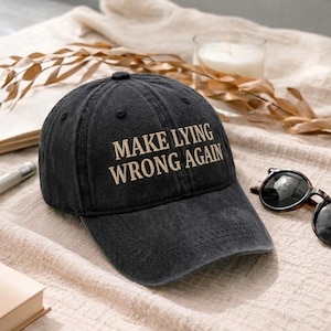 May include: A black baseball cap with the phrase "MAKE LYING WRONG AGAIN" embroidered in tan thread. The cap is sitting on a beige surface, with sunglasses and a candle in the background.