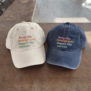 Puede incluir: Se muestran dos gorras de béisbol, una beige y otra azul marino. Ambas gorras tienen la frase bordada "keep the immigrants deport the racists" en varios colores. Están colocadas sobre una superficie de madera.