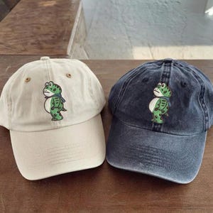 May include: Two baseball caps, one beige and one navy blue, are displayed on a wooden surface. Each cap features an embroidered cartoon frog wearing a scarf. The frog is green with white accents. The caps have a curved brim and adjustable strap.