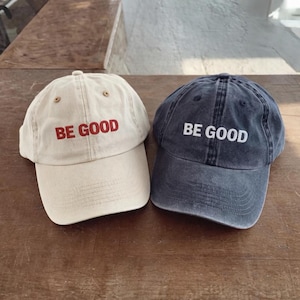 Puede incluir: Se muestran dos gorras de béisbol, una beige y otra azul oscuro. La gorra beige tiene el texto rojo "BE GOOD". La gorra azul oscuro tiene el texto blanco "BE GOOD". Ambas gorras tienen una visera curva y una correa ajustable.