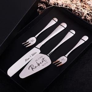 Può includere: Un set di coltello e server per torte argentati con due forchette, presentato in una scatola nera. Il coltello e il server sono incisi con nomi e una data. Le forchette e il coltello hanno una fascia nera decorativa vicino al manico.