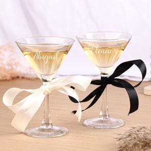 Può includere: Due bicchieri da martini trasparenti riempiti con un liquido giallo chiaro. Ogni bicchiere ha un nome inciso: "Abigail" ed "Eleanor". Un bicchiere è legato con un nastro avorio, l'altro con un nastro nero. I bicchieri sono su una superficie di legno chiaro.
